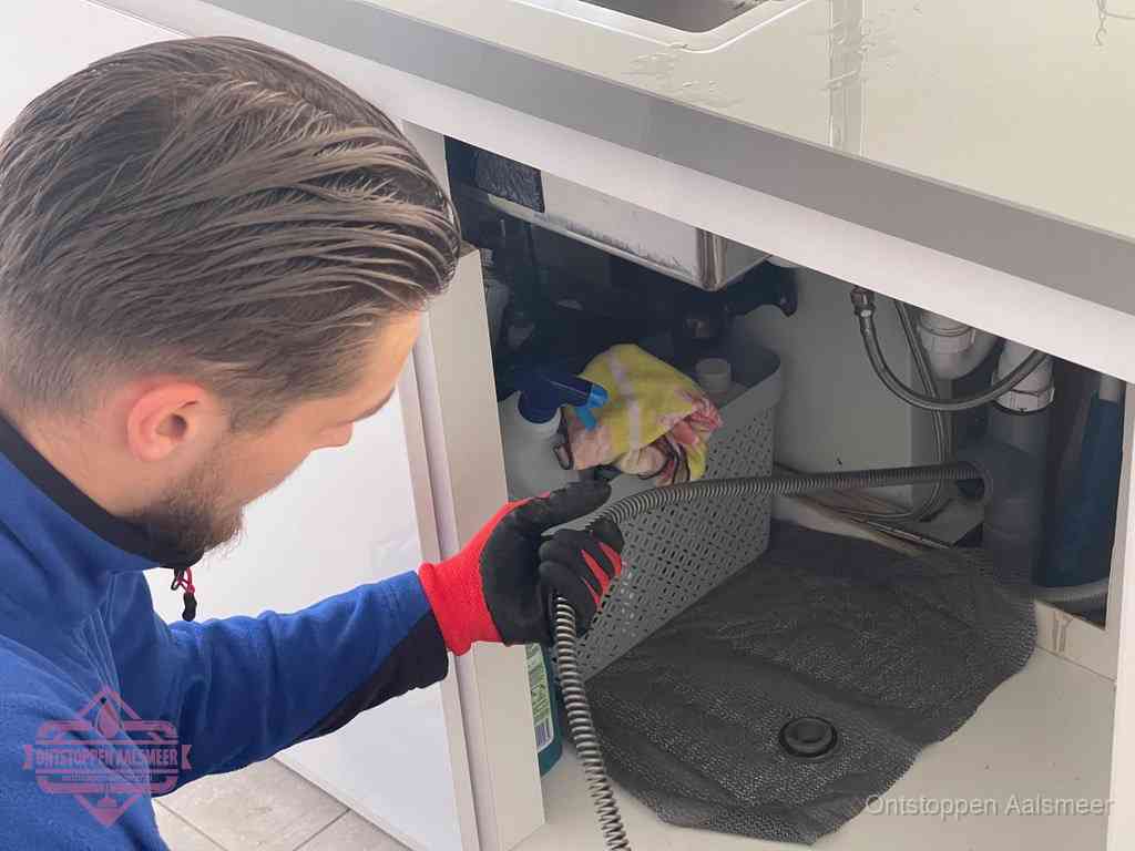 Een monteur voert een flexibele ontstoppingsveer in onder het keukenkastje, langs de sifon, om de gootsteen te ontstoppen in Aalsmeer