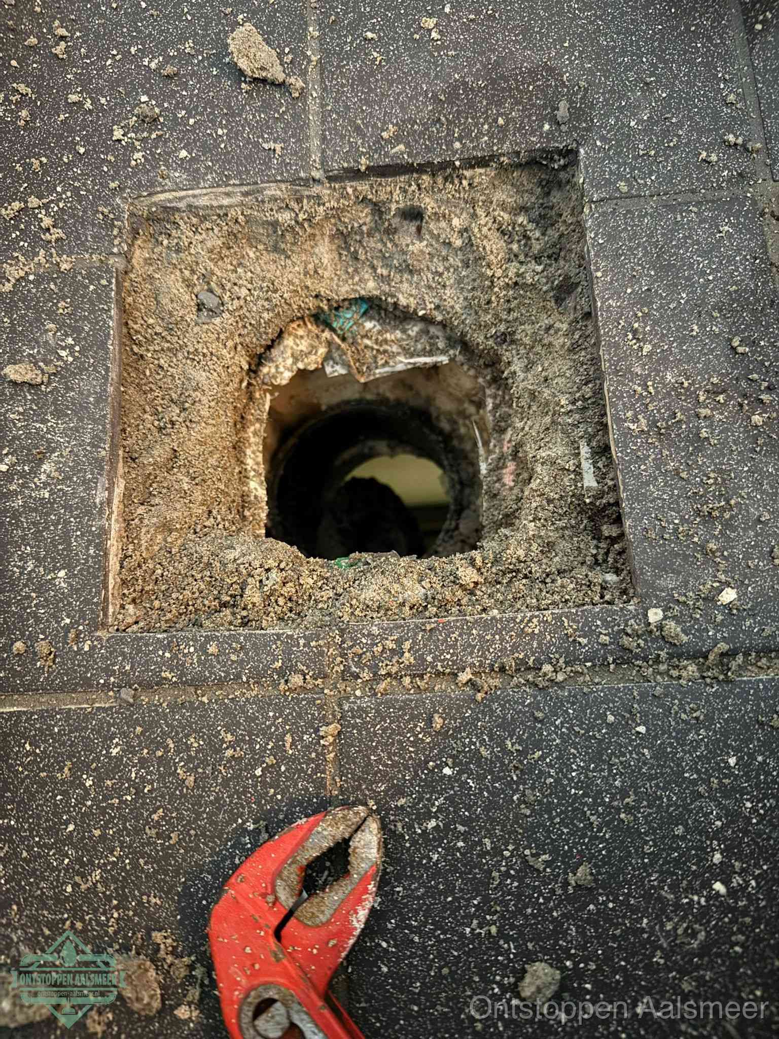 Een geopend rioolputje in een betontegelvloer met losgemaakt rooster en een zichtbare ronde afvoerbuis, het achterhalen waar de afvoerverstopping zit in Aalsmeer