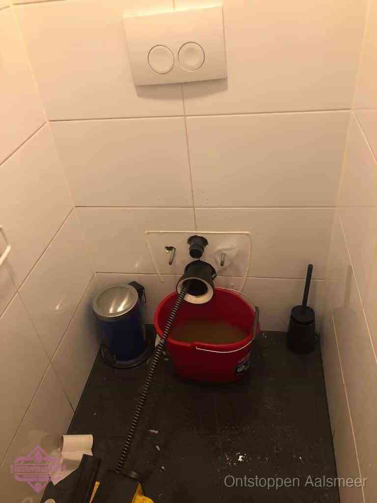 Een ontstopte wand-wc met verwijderde toiletpot waar een ontstoppingsveer in de wanduitgang verdwijnt, het achterhalen waar de verstopping zit in Aalsmeer