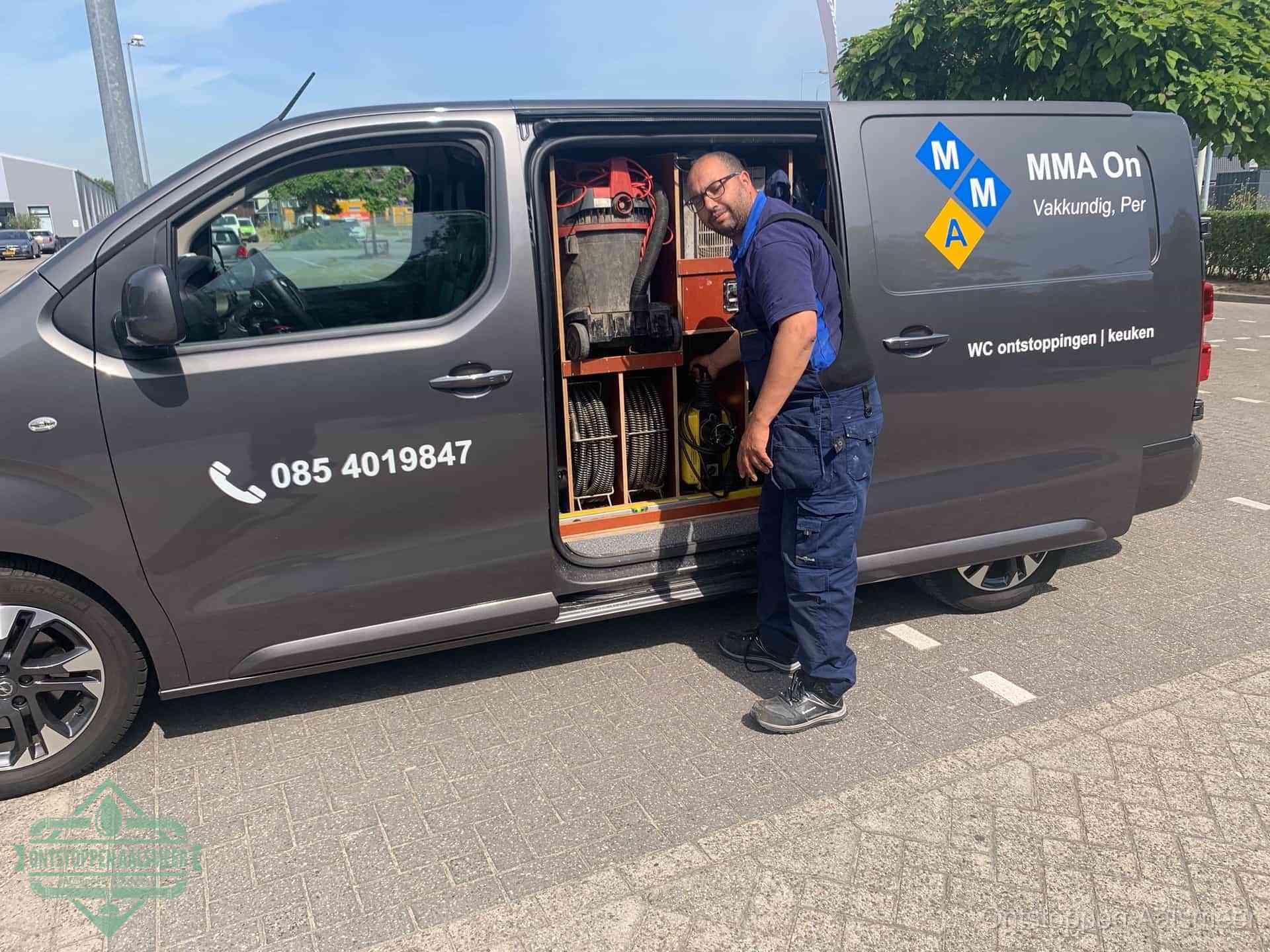 Een monteur bij de open schuifdeur van zijn bedrijfsbus op een parkeerplaats, klaar om de oorzaak van terugkerende afvoerverstoppingen aan te pakken in Aalsmeer