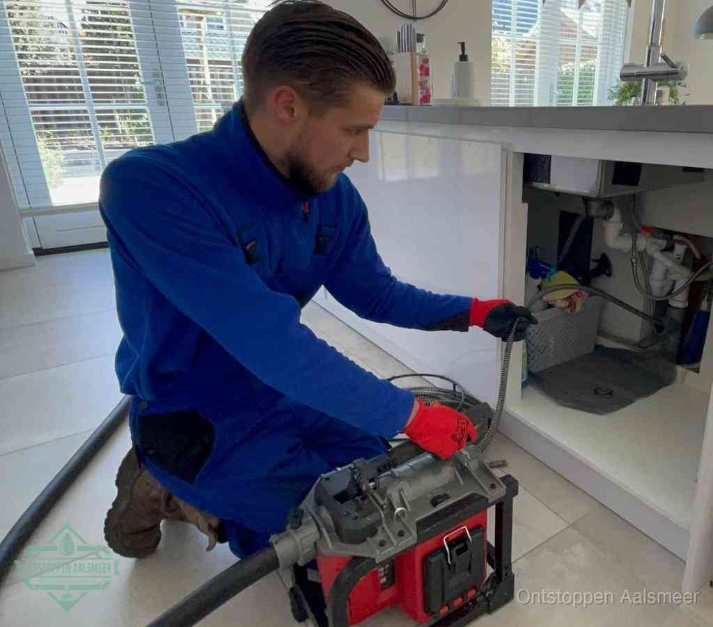 Een monteur met rode handschoenen knielt in een keuken en voert een elektrische ontstoppingsveer in tijdens het professioneel afvoer ontstoppen in Aalsmeer